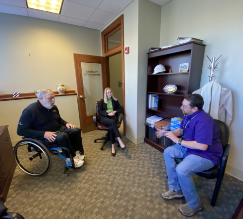 Man in wheelchair visits city staff inside city offices to talk about accessibility issues.