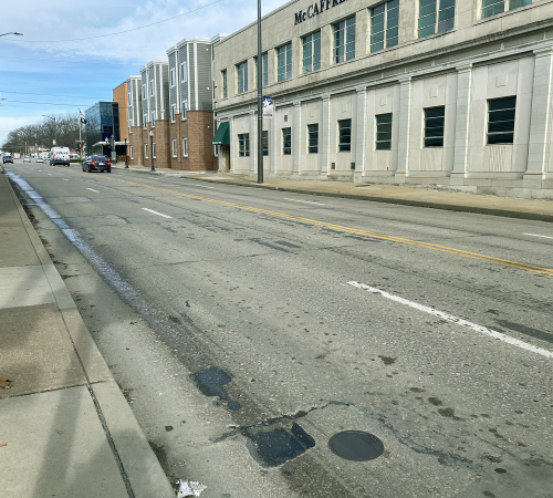 Photo of street next to buildings. The street shows patching and wear and tear.