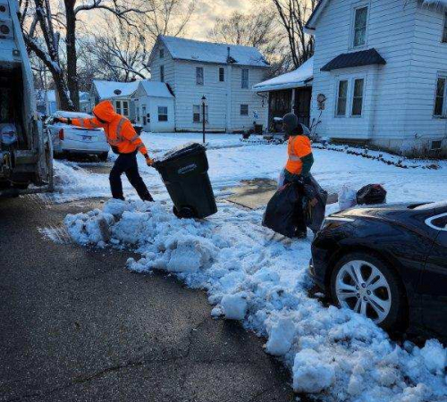 City crews in orange reflective jackets carrying trash bags through deep snow