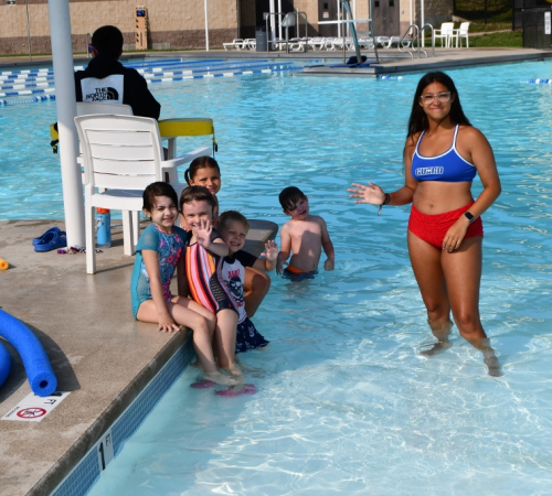 Woman in a bathing suit smiling and waving in a pool with a group of children