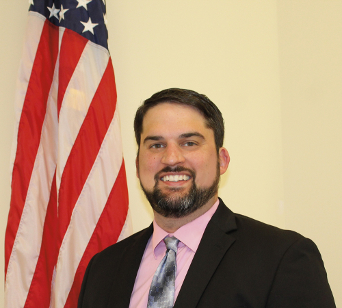 Image of man standing next to an American flag wearing a long necktie and black blazer