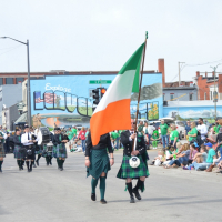 people walking in the middle of a parade street carrying an orange, white and green Irish flag