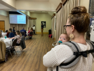 Mother and baby watch business presentation behind seated crowd in large room