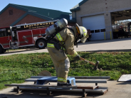 Firefighter practices using an axe in parking lot with fire station in the background