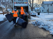 City crews in orange reflective jackets place trash bags into trash truck receptacle in neighborhood with deep snow