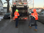 City crews in orange reflective jackets place trash bags into trash truck receptacle