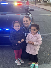 Woman in police uniform smiling with two small children in front of a police vehicle