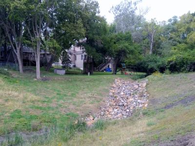 Photo of rip rap along new drainage ditch surrounded by green grass, trees and behind homes