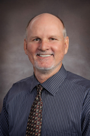 Man with no hair, grey moustache, blue button down shirt and tie