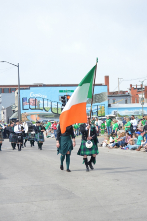 people walking in the middle of a parade street carrying an orange, white and green Irish flag
