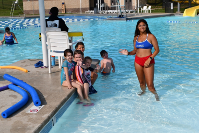 Woman in a bathing suit smiling and waving in a pool with a group of children