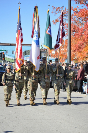 Honor Guard dressed in Army uniforms carry flags on a sunny street with orange trees turned for fall.