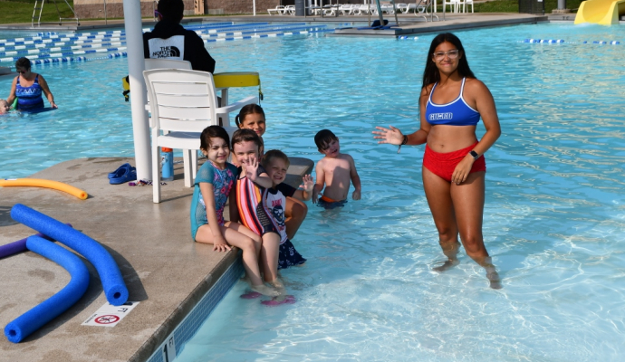 Woman in a bathing suit smiling and waving in a pool with a group of children